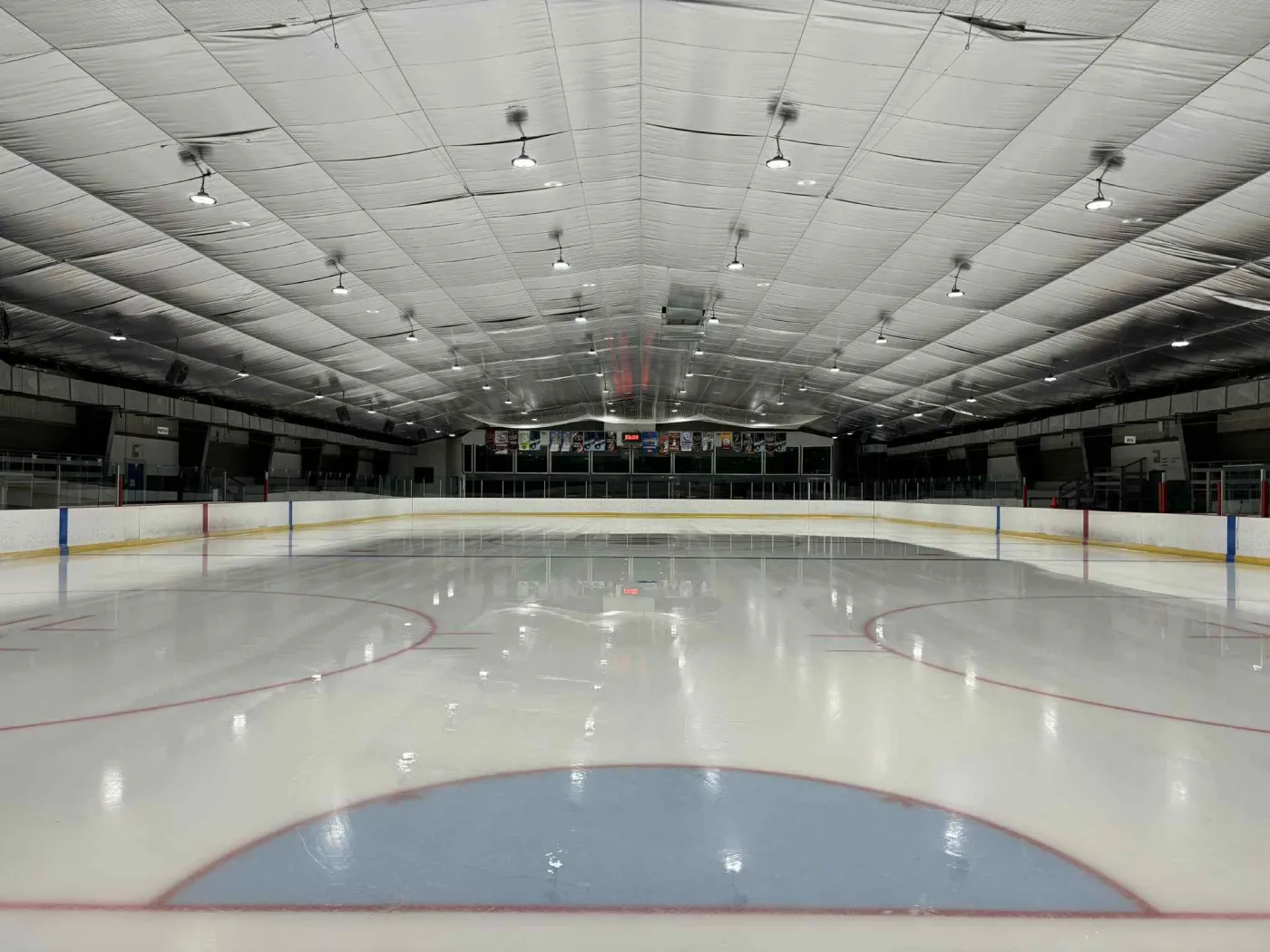 The empty ice sheet from the end of the arena, with red and blue lines, the boards, the scoreboard and the quilted ceiling. — Aréna de Métabetchouan-Lac-à-la-Croix