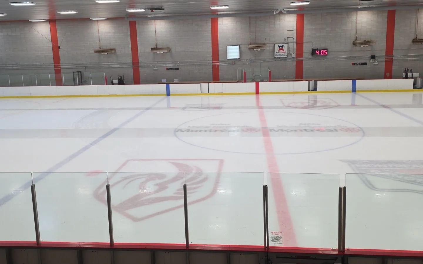 La pista vacía de Michel-Normandin desde las gradas, con el nombre de la arena pintado en el centro del hielo bajo pilares rojos y el reloj — Aréna Michel-Normandin