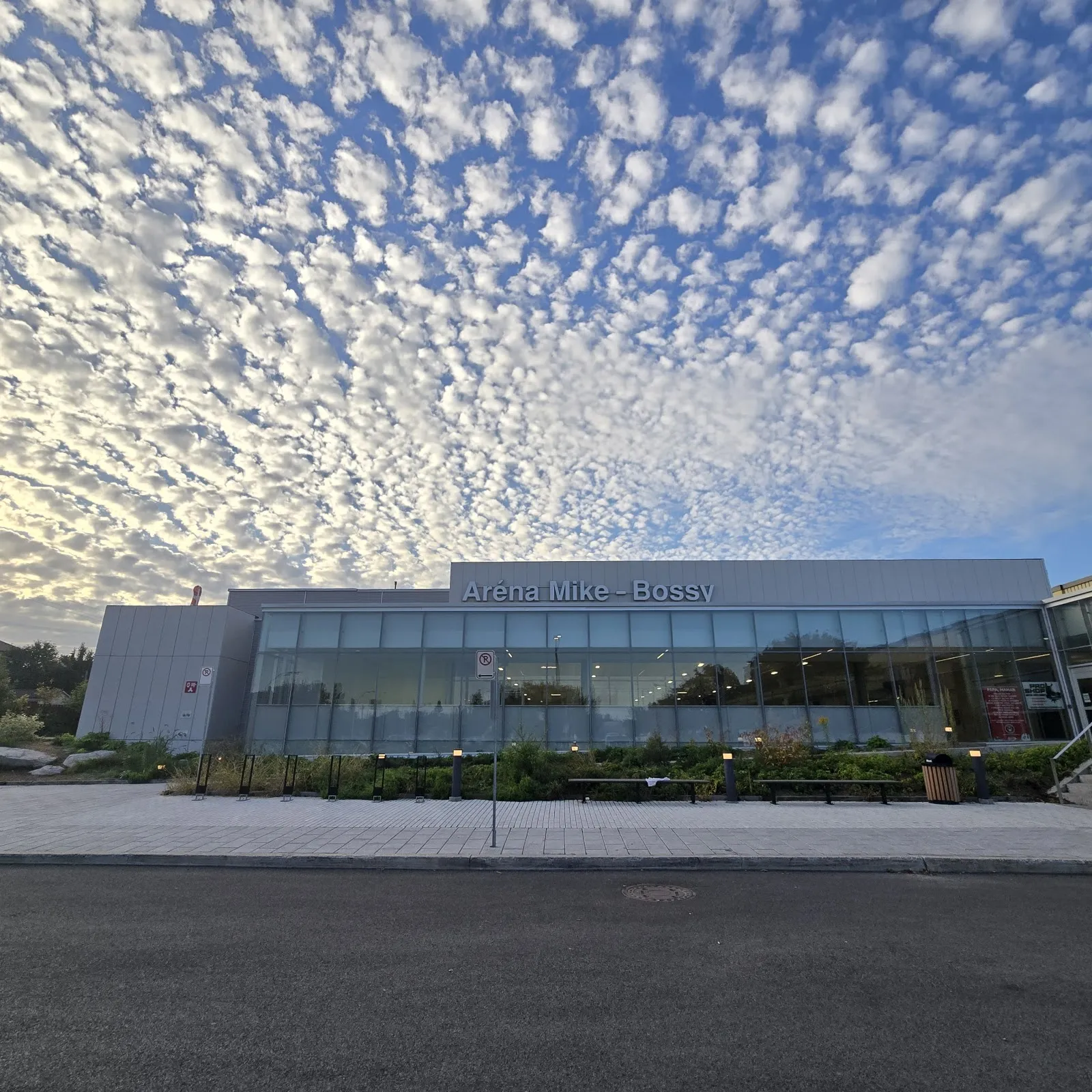 Le bâtiment vitré de l'Aréna Mike-Bossy avec son nom en lettres métalliques au-dessus du long mur de fenêtres, une place pavée devant et un ciel de petits nuages au crépuscule. — Aréna Mike-Bossy (Laval)