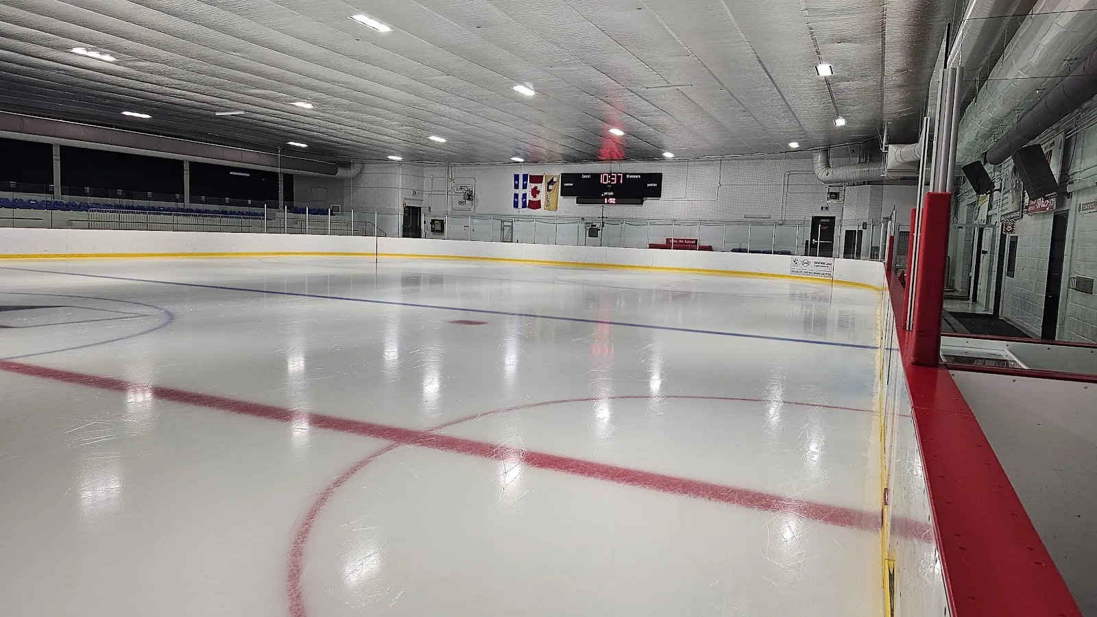 Un coin de la surface de glace vide vu par-dessus les bandes rouges, avec la ligne rouge centrale et un cercle de mise au jeu, la plinthe jaune et le tableau indicateur au fond de la salle. — Aréna Mike-Bossy (Laval)