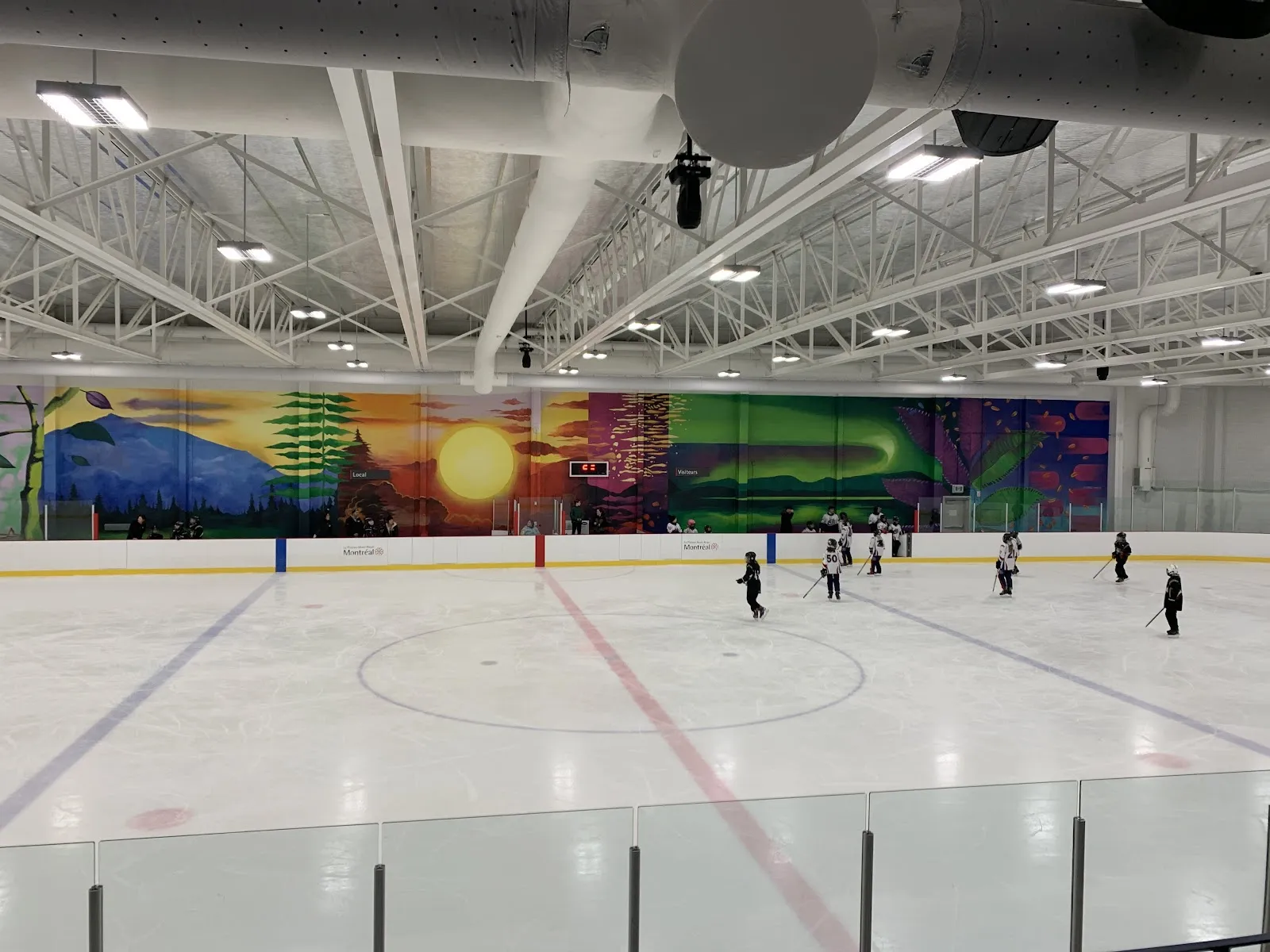 La surface de glace complète vue de la bande pendant une séance de hockey mineur, avec une murale de paysage colorée au mur du fond et l’image de marque « Montréal » sur les bandes. — Aréna Mont-Royal
