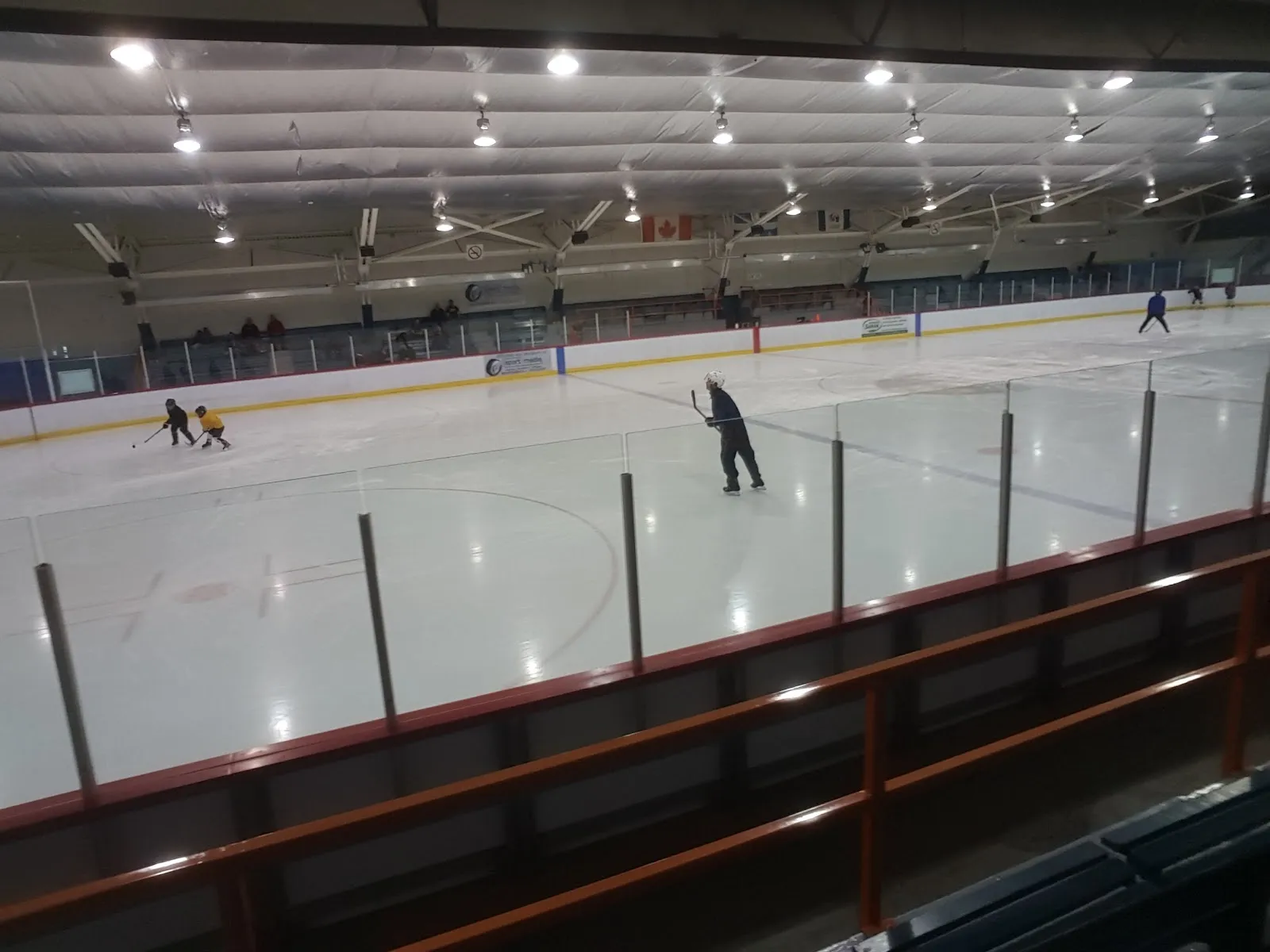 La pista de hielo vista a través del cristal desde las gradas, con unos pocos patinadores y dos jóvenes jugadores pasándose un disco, las vallas con sus paneles de patrocinadores, la bandera de Canadá y el techo aislado encima. — Aréna municipal de Saint-Jean-sur-Richelieu