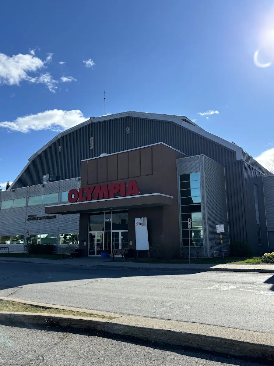The arena's entrance under the red OLYMPIA lettering, with the curved metal roof of the rink hall behind it. — Aréna Olympia
