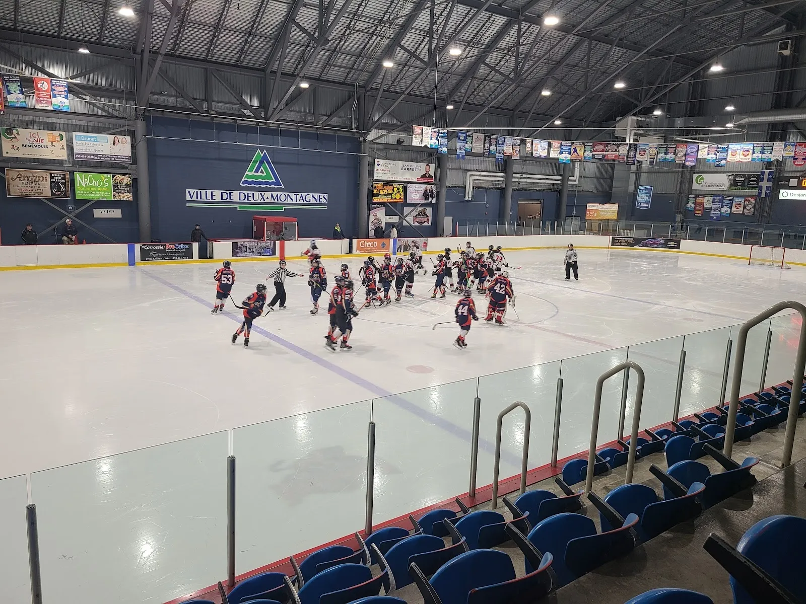A team on the ice in front of the blue seats, players spread across a wide sheet beneath a dark steel roof and a team banner wall. — Aréna Olympia