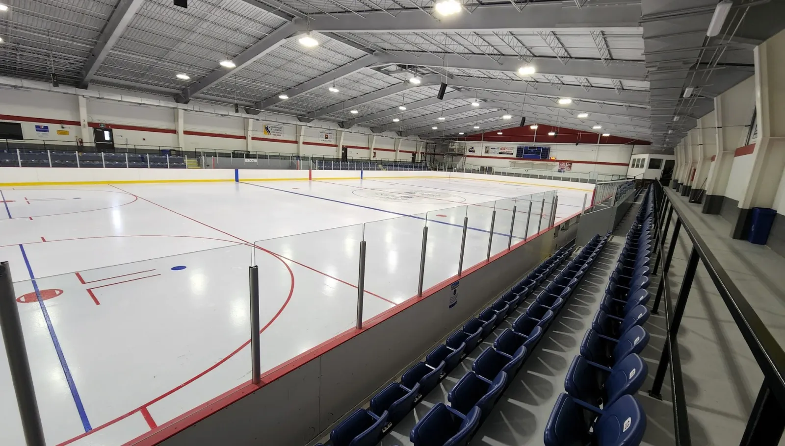 La patinoire vue d’une rangée surélevée de sièges bleus dans le coin, sous une charpente d’acier grise, avec des bandes blanches sur une glace fraîche. — Aréna Patrick-Poulin