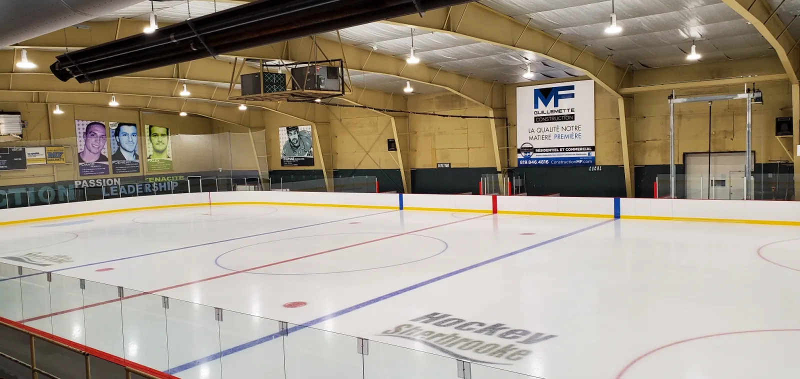 The empty rink under a wooden arched roof, with player portrait banners on the end wall and Hockey Sherbrooke painted on the ice. — Aréna Philippe-Bergeron