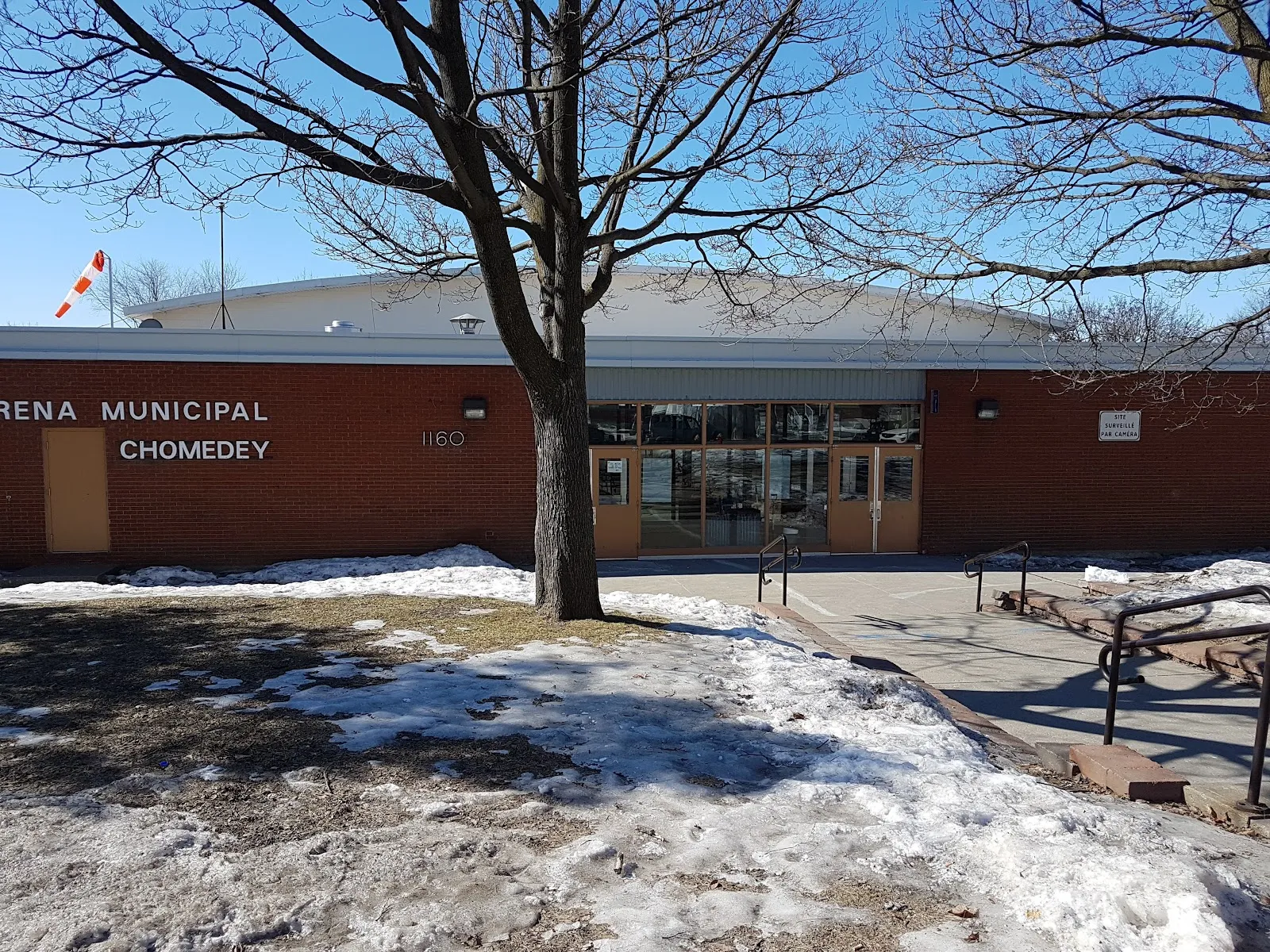 The red-brick front of the arena with the civic number 1160 above its glass doors, its municipal sign reading Chomedey, a bare tree and patches of snow in front. — Aréna Pierre-Creamer (Laval)