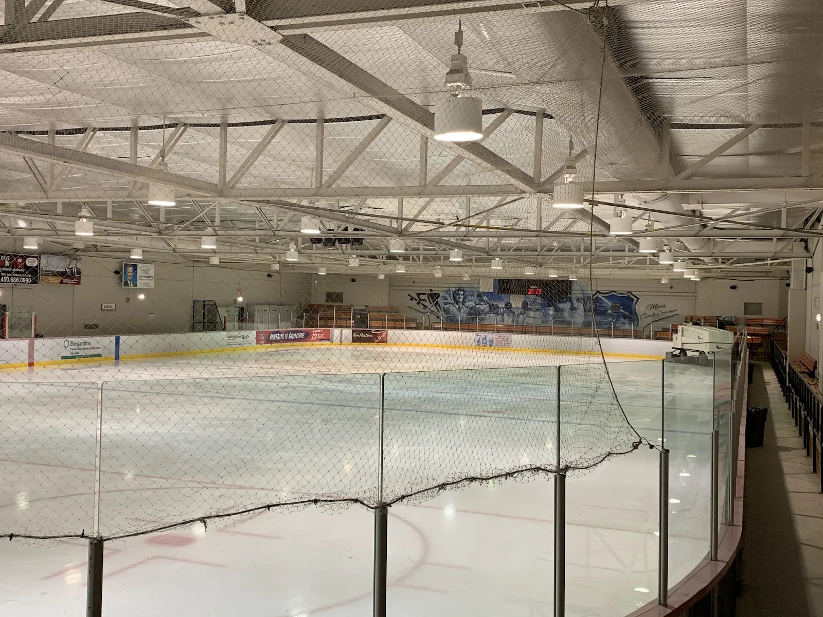 The ice sheet seen from the walkway above the boards through protective netting, with the steel roof trusses, the far-side stands and boards with sponsor panels, and no one on the ice. — Aréna Pierre-Creamer (Laval)