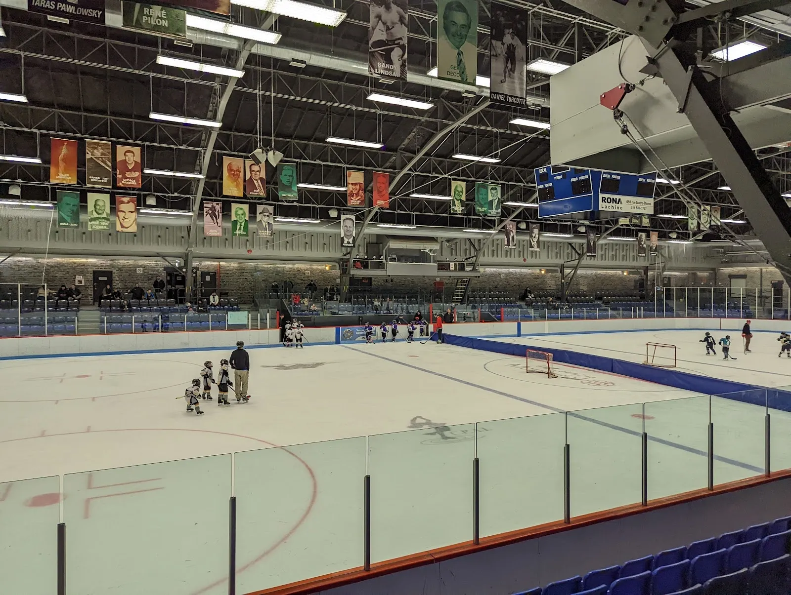 A sheet from the stands with banners above the far wall and a goal in the distance, a few skaters and children at the boards under a high steel roof. — Aréna Pierre « Pete » Morin