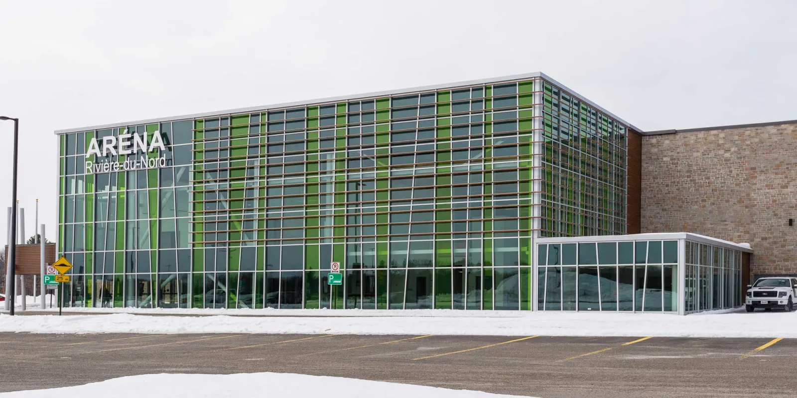 La fachada de vidrio y paneles verdes de la arena, con las letras de Aréna Rivière-du-Nord, junto a un ala de ladrillo y un estacionamiento nevado. — Aréna régional de la Rivière-du-Nord