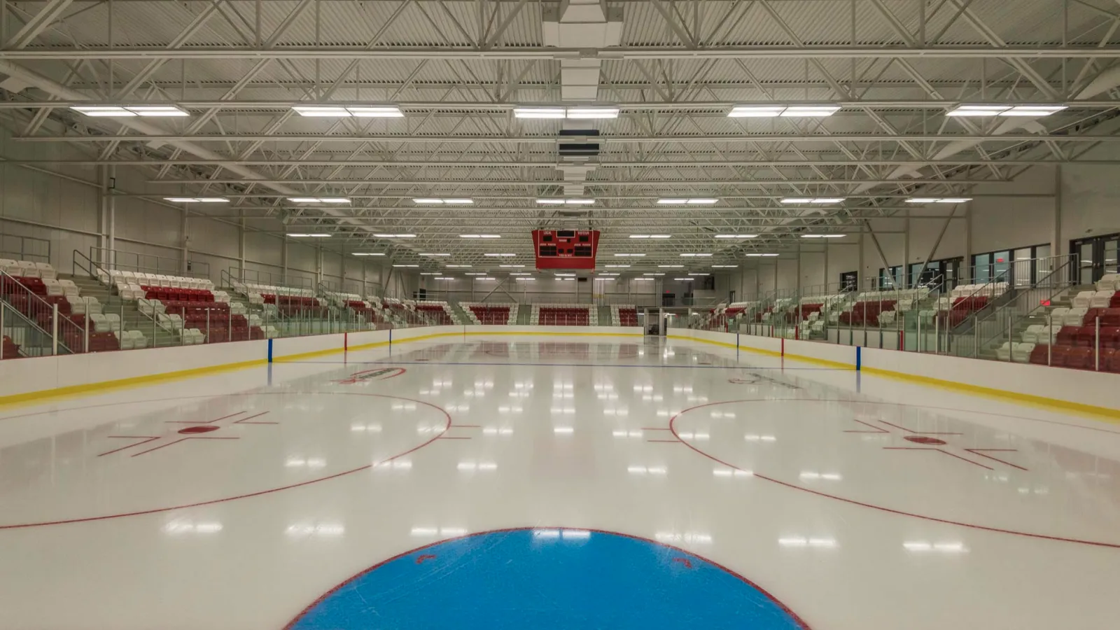 La pista principal vacía a nivel del hielo desde el centro, con asientos rojos y blancos a ambos lados y un marcador colgado en el extremo opuesto. — Aréna régional de la Rivière-du-Nord