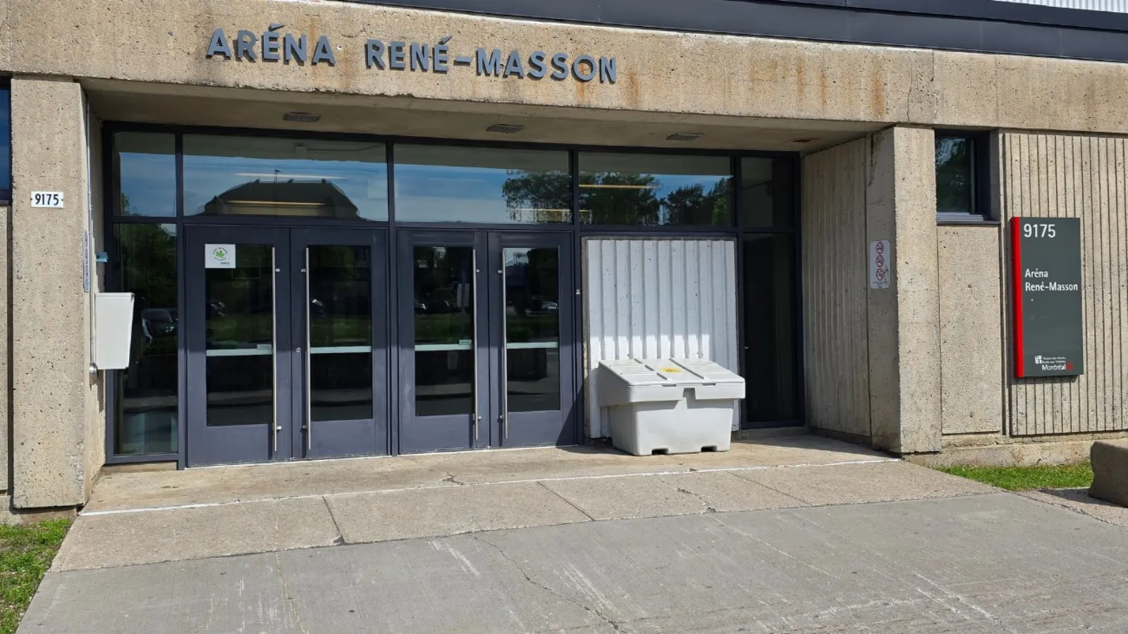 L’entrée principale de l’Aréna René-Masson, avec le nom en lettres sur la façade de béton au-dessus des portes vitrées et une enseigne 9175 à côté. — Aréna René-Masson