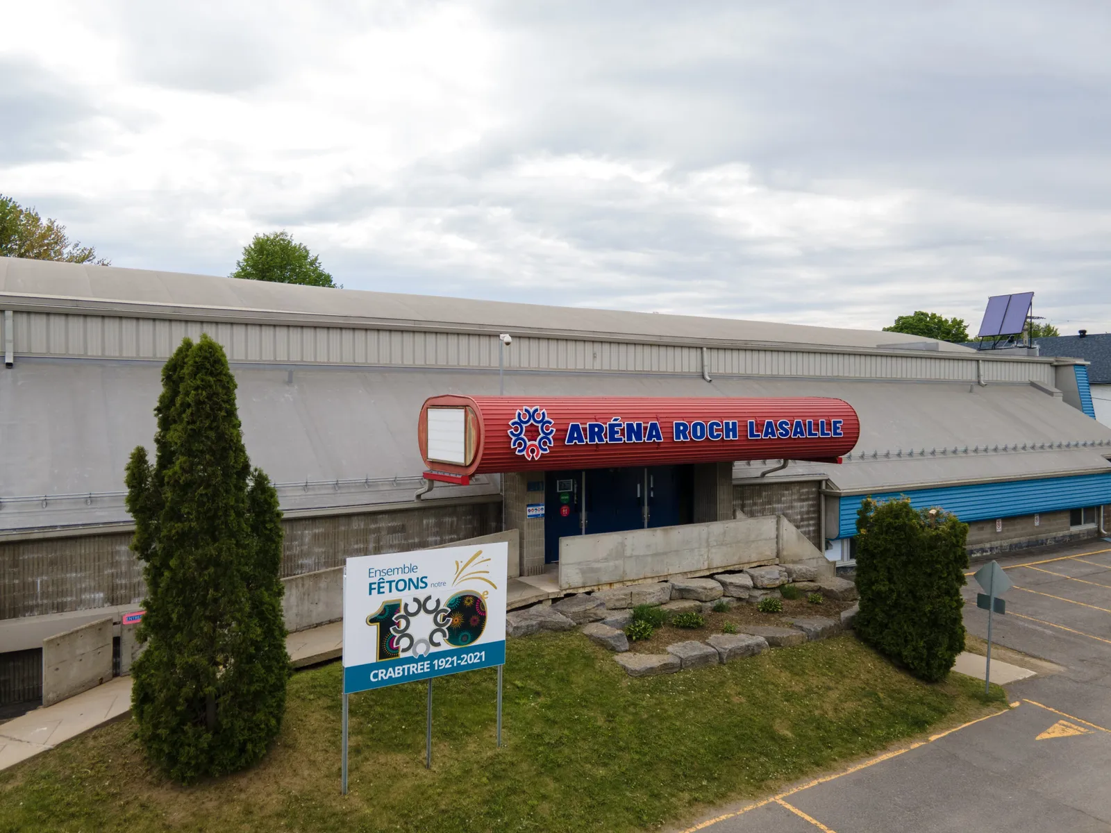 The Aréna Roch-Lasalle in Crabtree seen from the street: a long low building with a red and blue sign above the entrance and a conifer in front. — Aréna Roch-Lasalle