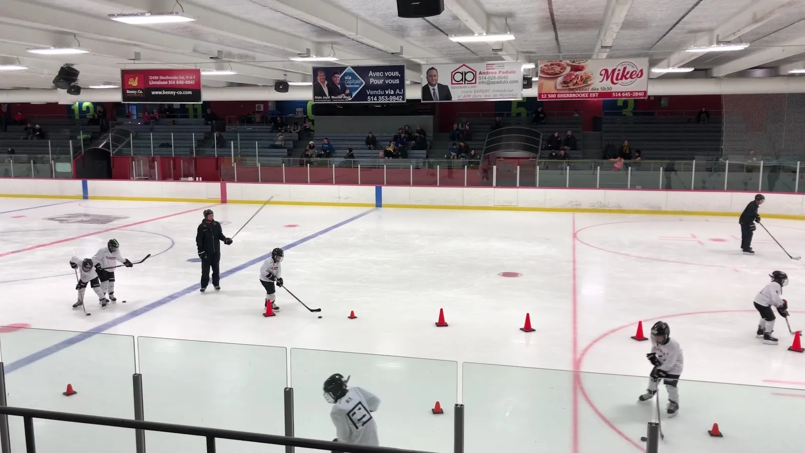 Un entraînement avec des cônes répartis sur une grande surface de glace, des joueurs et des entraîneurs sur la glace sous un long plafond bas. — Aréna Rodrigue-Gilbert