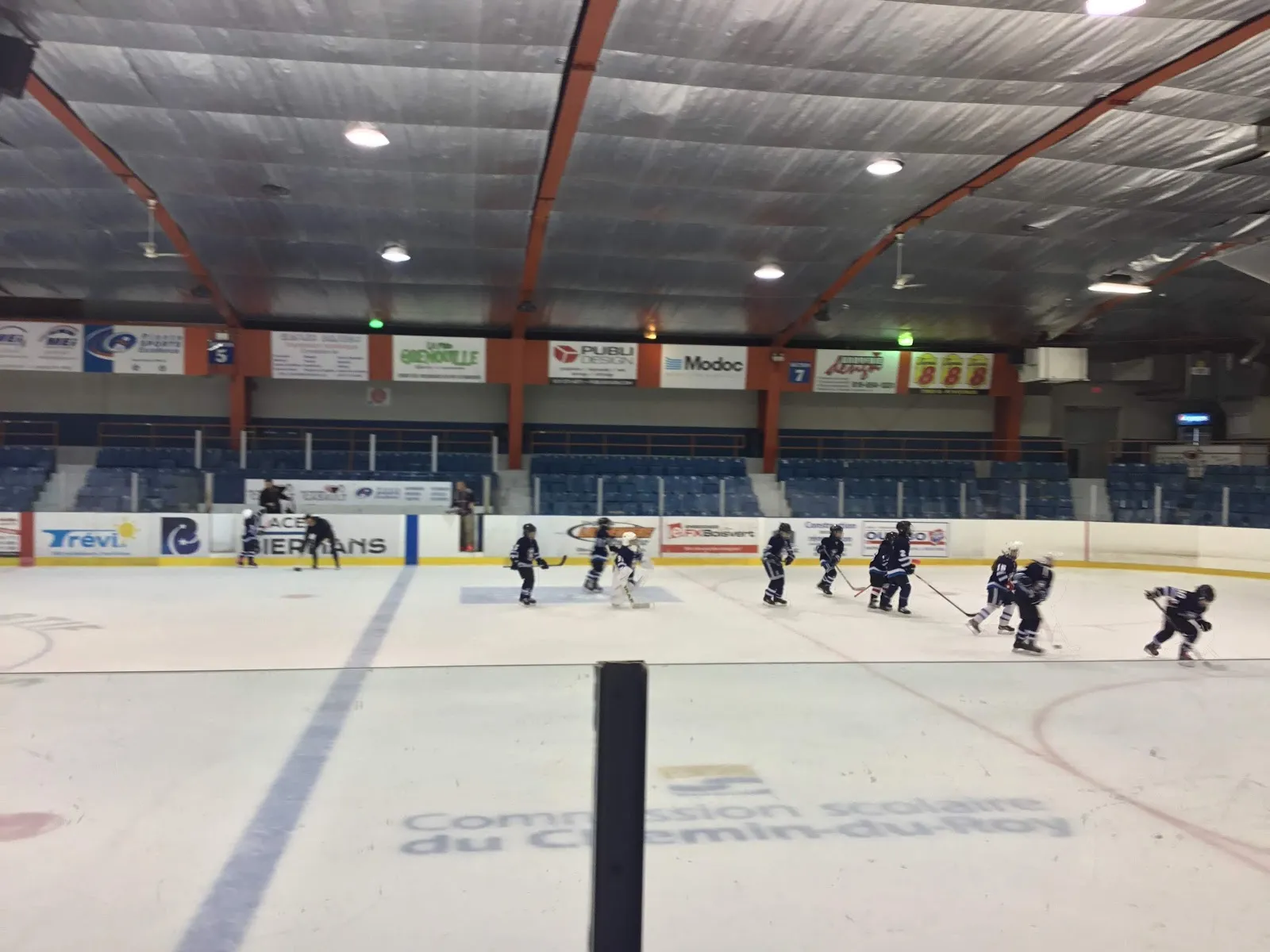 Skaters practising on the ice at the Aréna Roland-Rheault, with the boards and sponsor signs behind — Aréna Roland-Rheault
