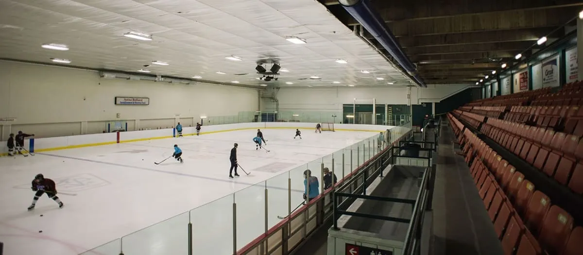An empty-looking sheet seen from the stands, a few skaters spread across the ice beside a long run of orange seats and a glass-topped rail. — Aréna Rolland