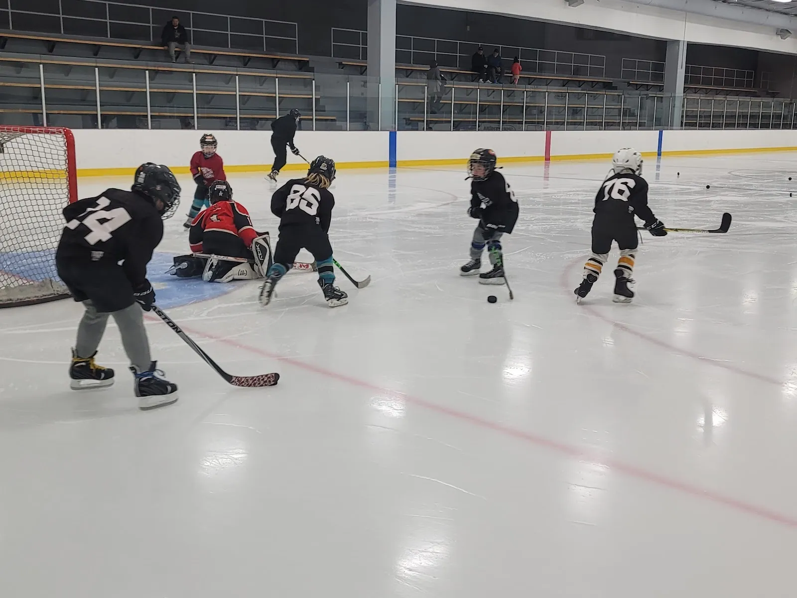 De jeunes joueurs à l’entraînement près du filet sur une glace d’un blanc éclatant, une surface polie par la surfaceuse avec des lignes rouges et bleues et la rampe des gradins le long du côté opposé. — Aréna Saint-Charles