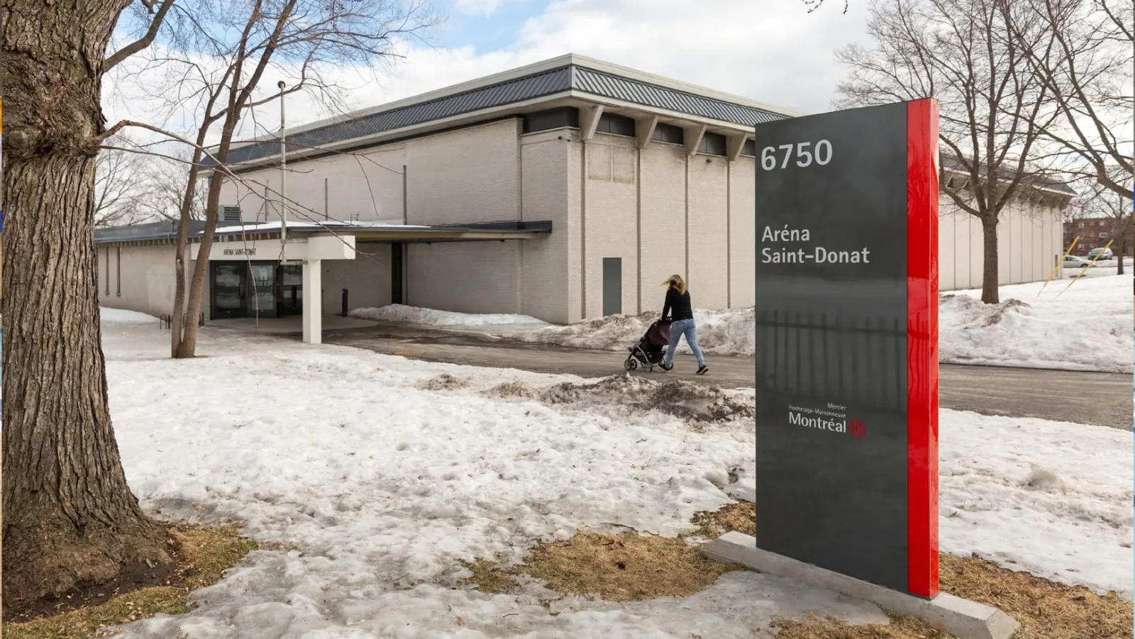 L’entrée principale du bâtiment d’aréna en brique crème avec une marquise au-dessus des portes; une enseigne municipale autoportante devant indique « 6750 Aréna Saint-Donat » avec le logo de Montréal / Mercier–Hochelaga-Maisonneuve, et « ARÉNA SAINT-DONAT » est inscrit sur la marquise. — Aréna Saint-Donat