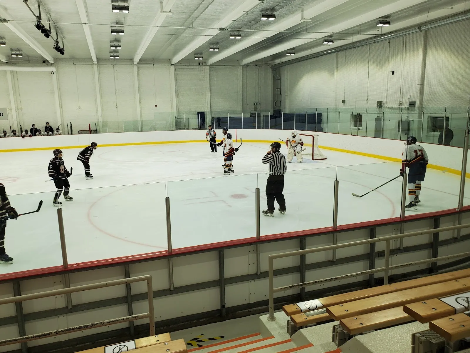 Un match vu des gradins par-dessus un banc de bois, des joueurs répartis sur la glace sous un plafond pâle, avec un mur gris derrière. — Aréna Saint-Donat