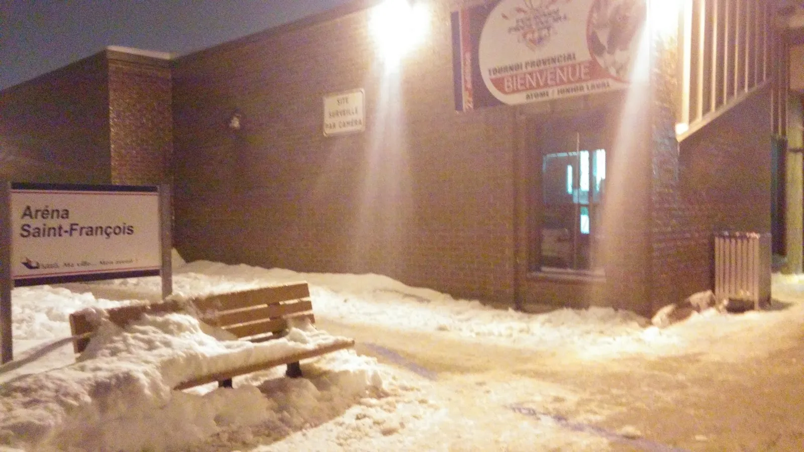 La esquina de entrada del Aréna Saint-François de noche: el nombre en el letrero azul de la calle junto a un banco rodeado de nieve, con una pancarta de bienvenida y una puerta acristalada en la pared de ladrillo. — Aréna Saint-François (Laval)