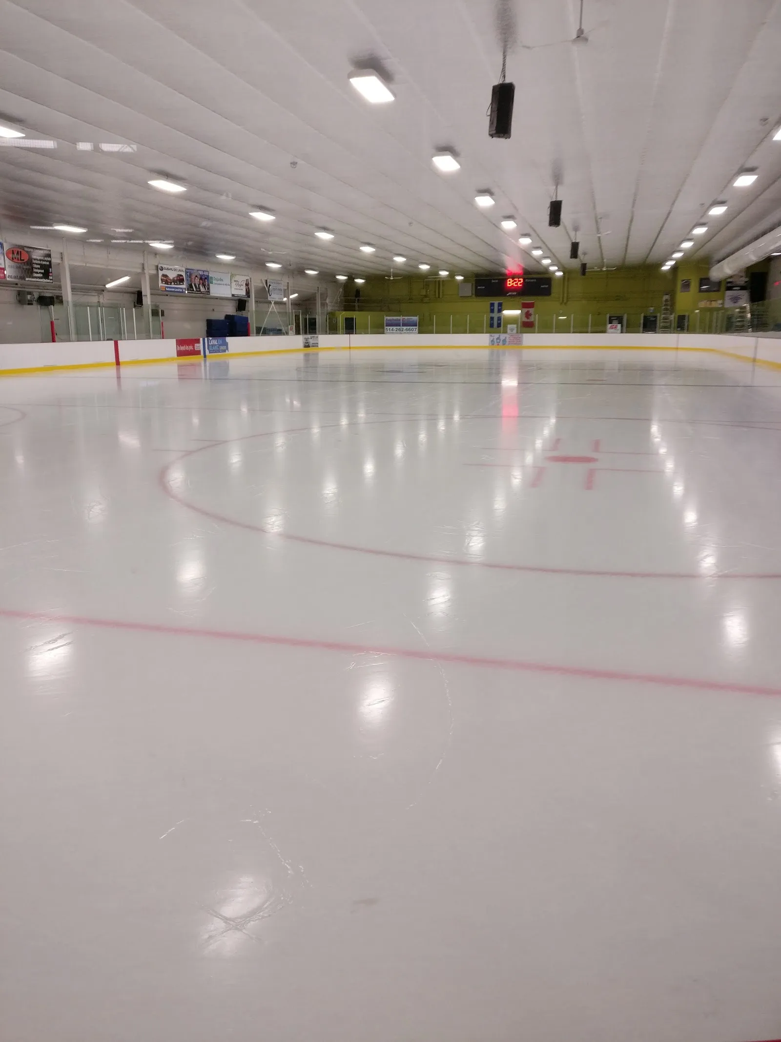 La pista vacía a ras de hielo, con la línea central roja y el círculo de salida en primer plano, el marcador, las pancartas retiradas y la pared verde del fondo, y el techo blanco abovedado. — Aréna Saint-François (Laval)