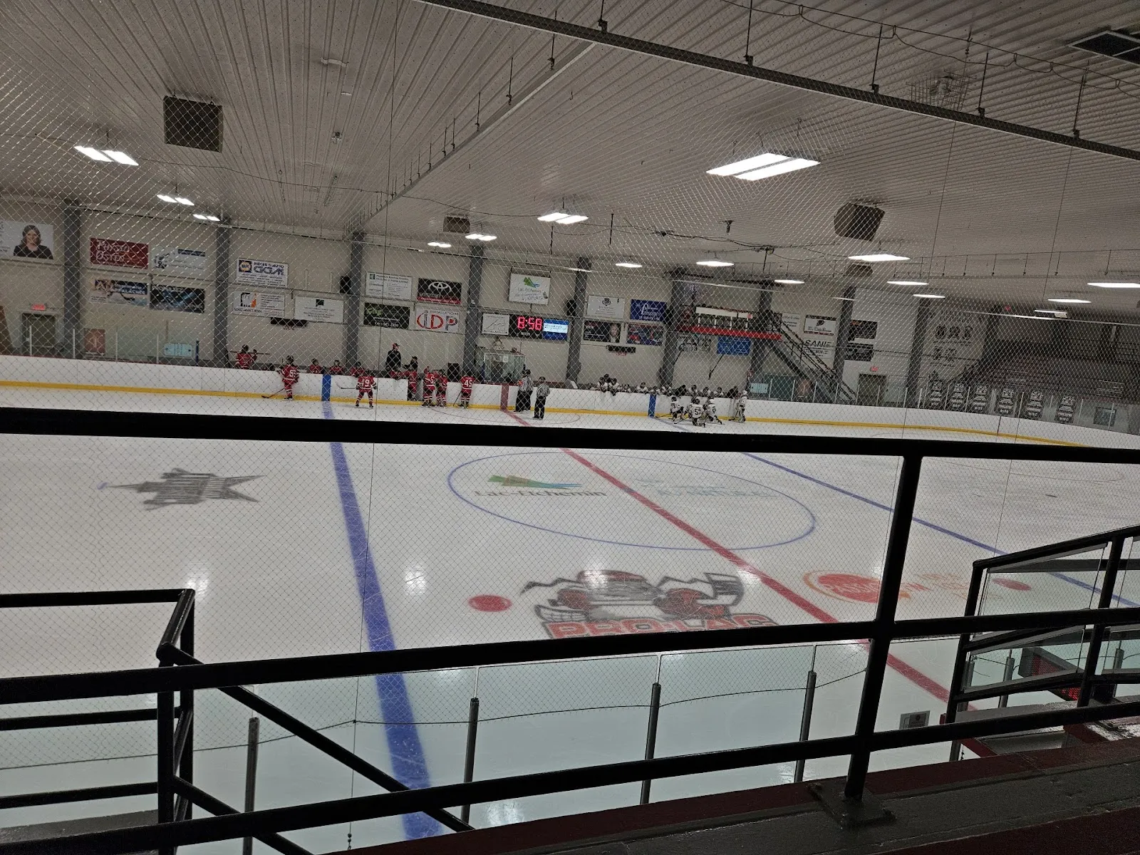Toda la pista de hielo vista desde detrás de un barandal en la parte alta de las gradas, con el logotipo del centro del hielo, letreros de patrocinadores a lo largo de las bardas y una hilera de luces bajo el techo. — Aréna Simon-Nolet