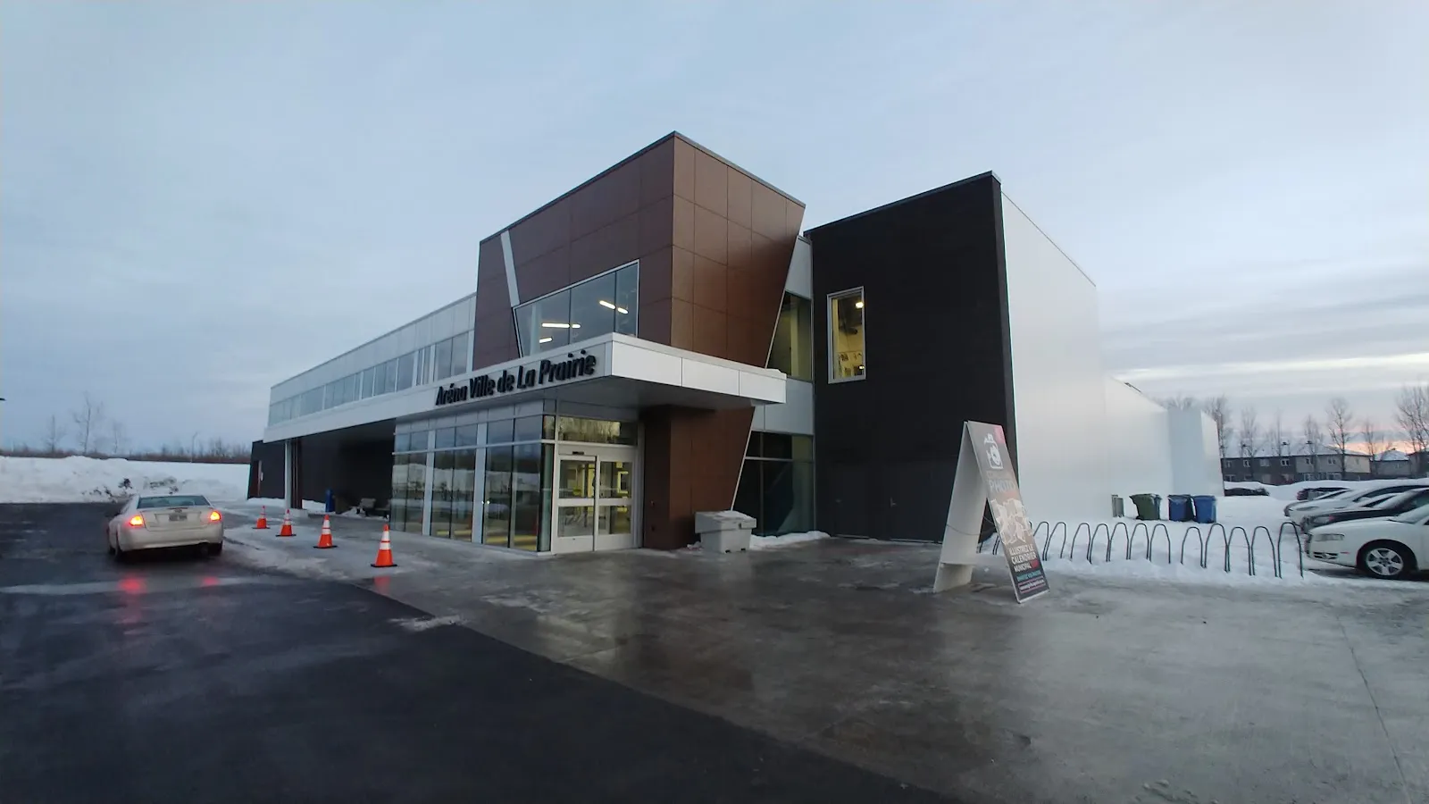 The modern glass and wood-panel entrance of the arena at dusk, signed Aréna Ville de La Prairie, with a dark grey block beside it. — Aréna Ville de La Prairie
