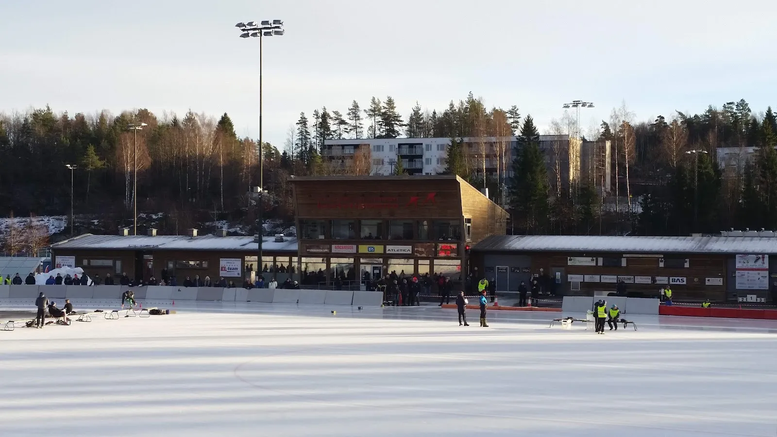 The brick-and-timber clubhouse and floodlights of the Arendal oval seen across the ice on a winter day. — Arendal kunstisbane (Skøytebanen)