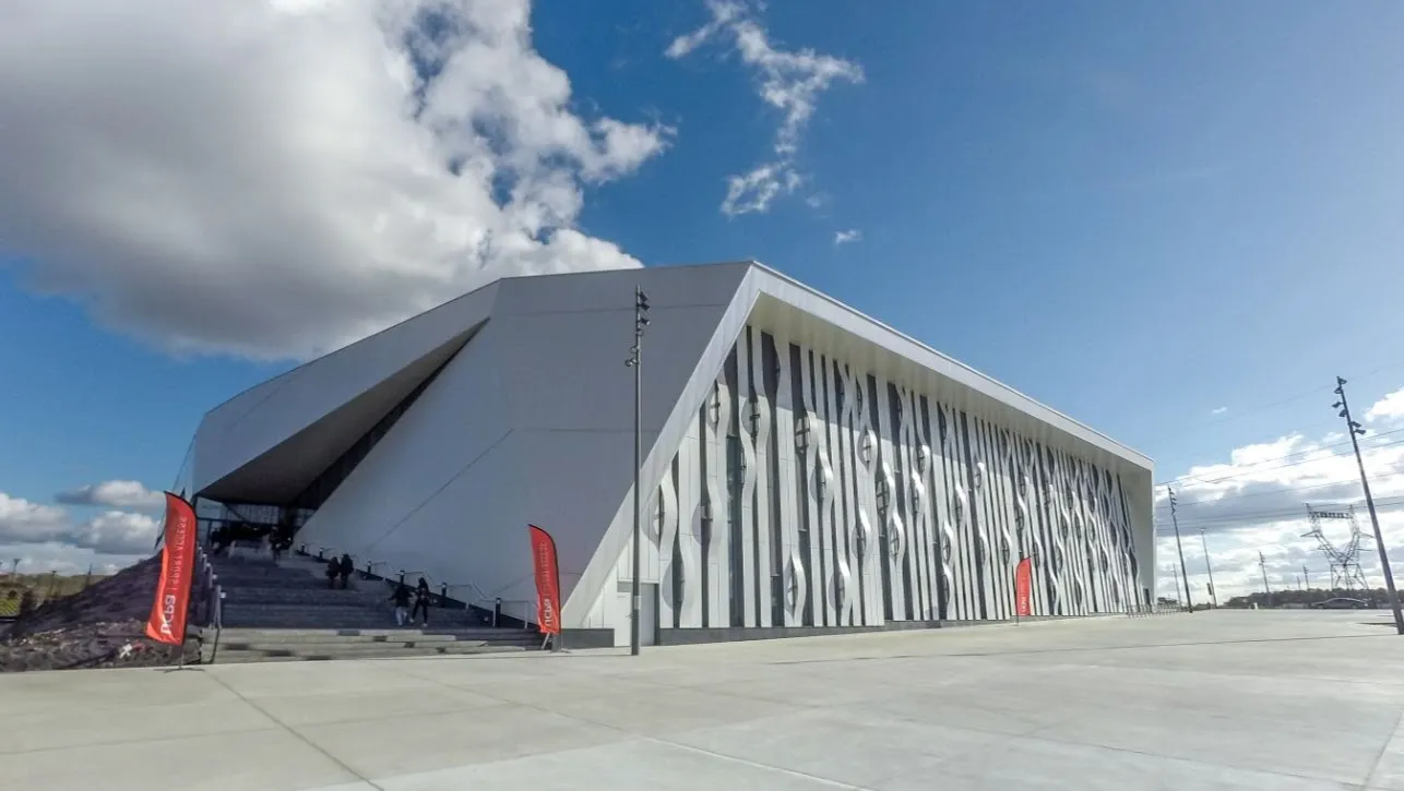 The faceted white Aren'Ice building seen from the forecourt under a blue sky, with a dark patterned cladding panel on the side wall and people at the entrance steps. — Aren'Ice Cergy-Pontoise