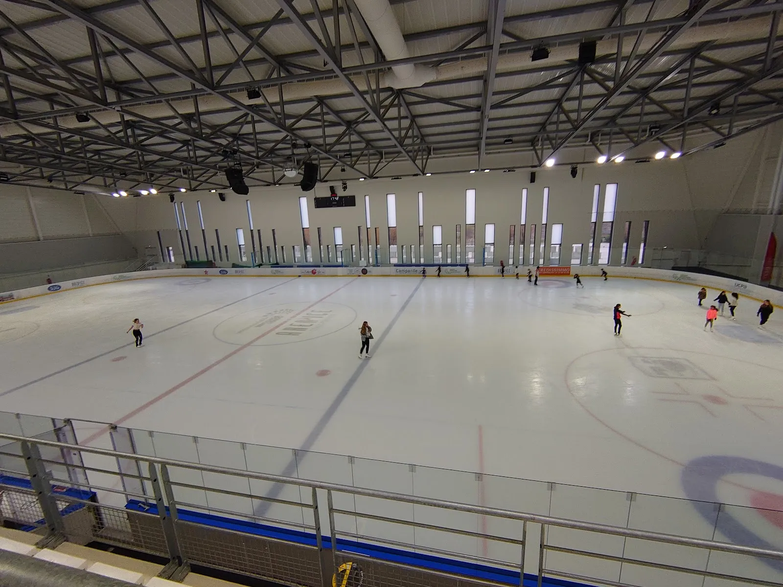 One of the two rinks seen from the stands at a public session: the full sheet of ice with a few skaters, long boards along the side and the steel roof trusses above. — Aren'Ice Cergy-Pontoise
