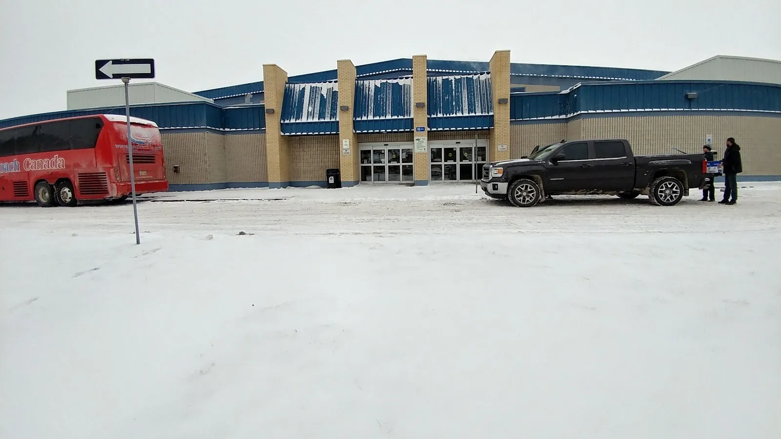 La entrada de la arena entre pilares de ladrillo beige, bajo franjas de techo metálico azul, con un estacionamiento nevado y un autobús estacionado al frente. — Argyle Arena