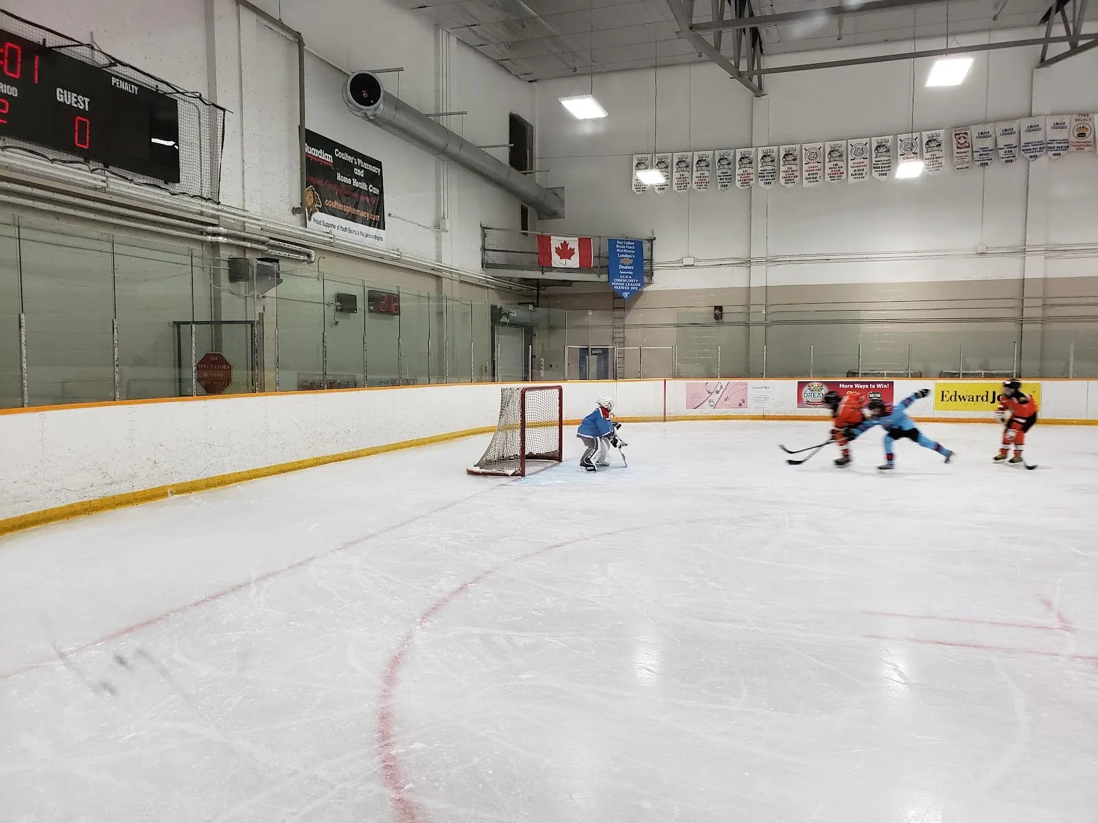 Un partido en juego en un extremo de una pista amplia visto por encima de las vallas, con jugadores alrededor de la portería bajo un techo de acero con una bandera de Canadá y un reloj de marcador. — Argyle Arena