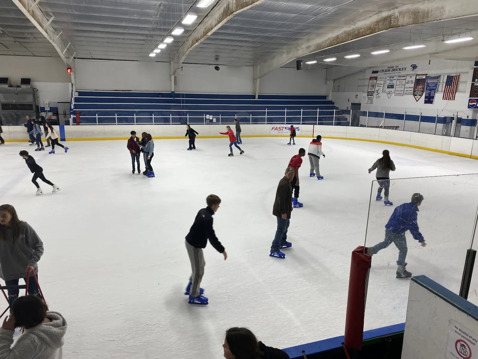 A public ice session on the white sheet with skaters spread across the ice, boards and a wall of banners behind them under a low ceiling. — Arkansas Skatium