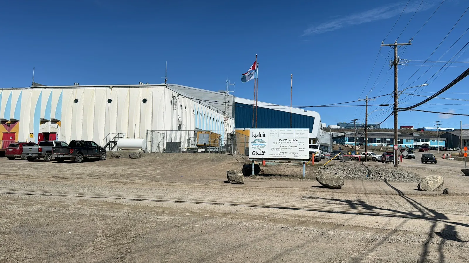 The pale metal Arnaitok Arena building with its roadside sign and a flagpole over a gravel lot. — Arnaitok Arena