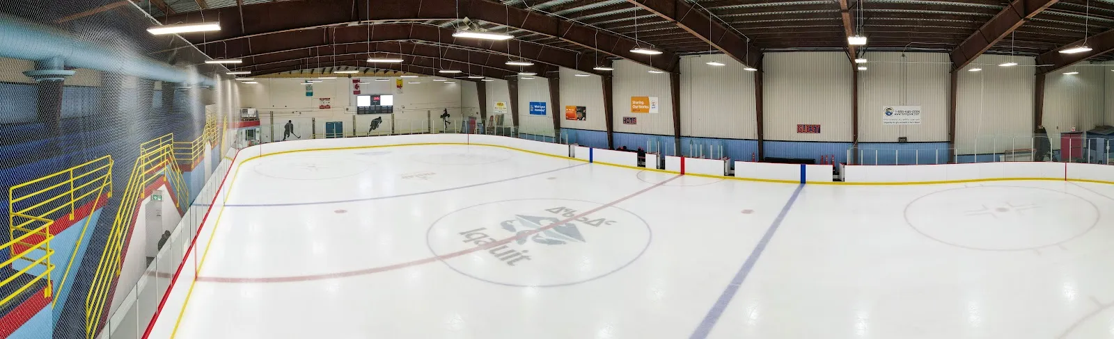 The empty Arnaitok Arena ice with centre-ice logo, orange boards and the steel roof overhead. — Arnaitok Arena