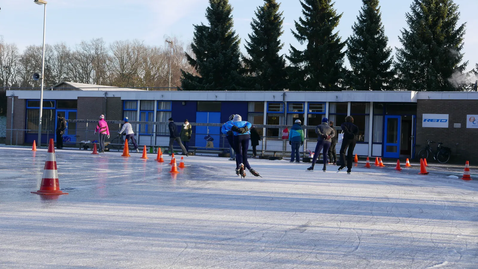 Schaatsers op afstand op de natuurijsbaan van Sportpark 't Cranevelt, met het clubhuis met blauwe kozijnen en een rij hoge sparren achter de pylonen. – Arnhemsche IJsclub Thialf (Arnhem)