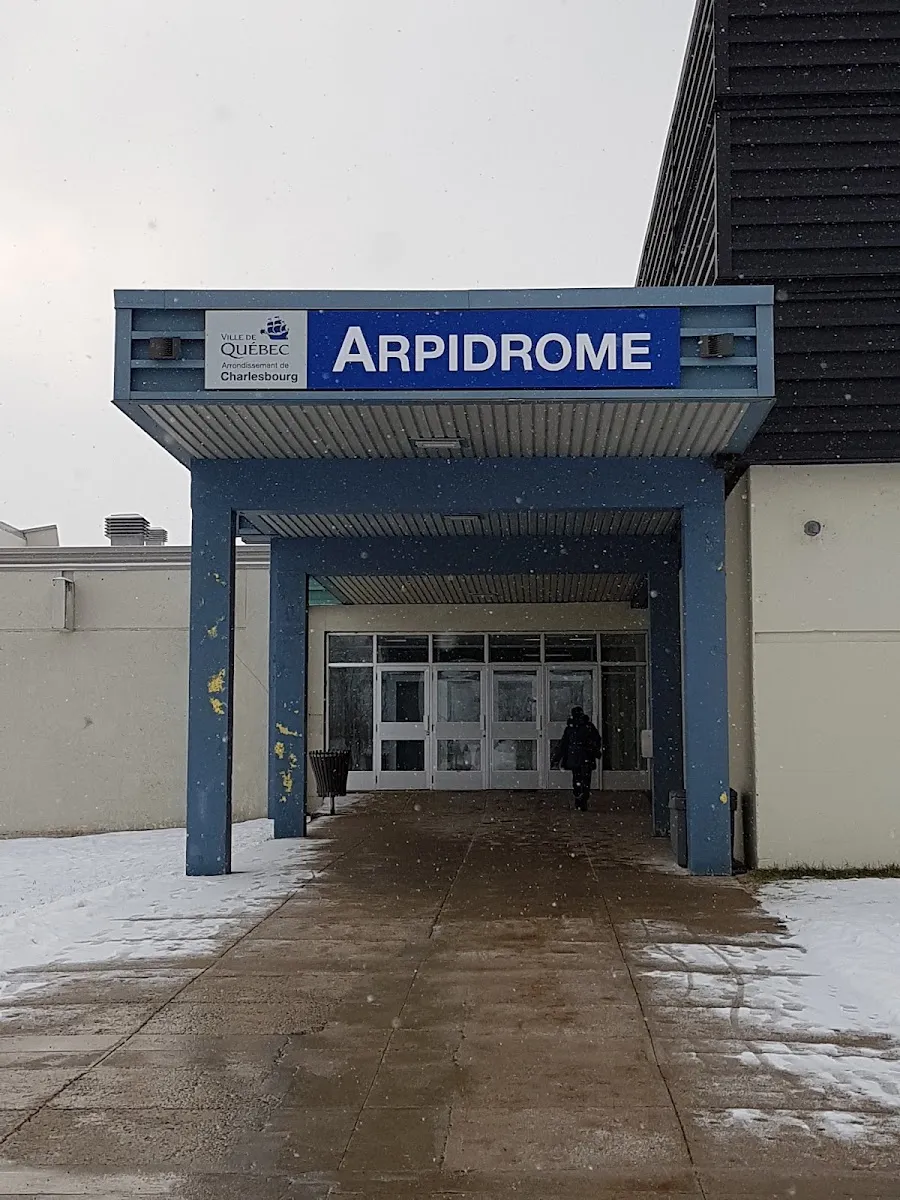 L'entrée à auvent bleu de l'Arpidrome, avec son nom sur une enseigne bleue à côté de l'écusson de la Ville de Québec et une allée saupoudrée de neige menant aux portes vitrées. — Arpidrome