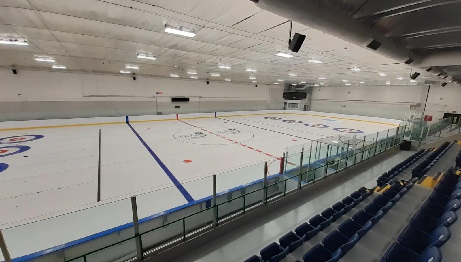 Une vue en longueur de la glace vide depuis les gradins, avec des sièges bleus sur le côté, des cercles de style curling peints dans la glace et un plafond blanc. — Arpidrome