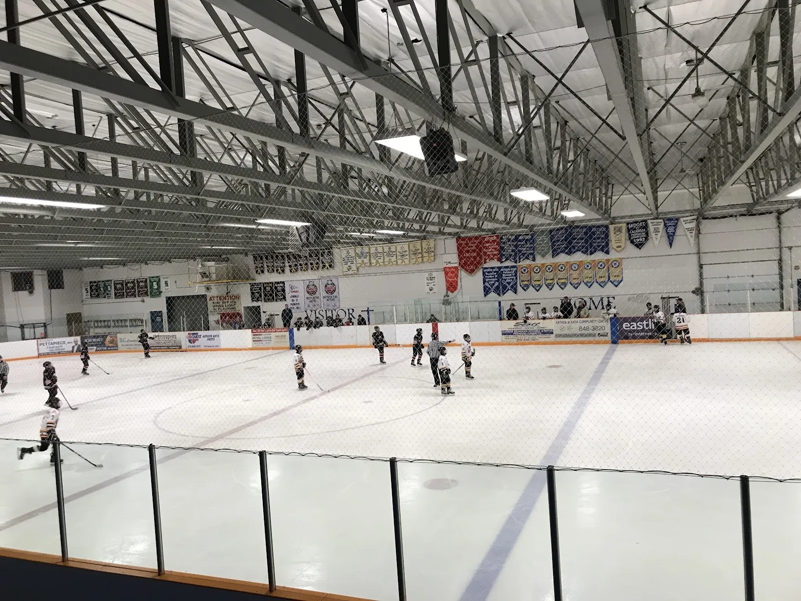 A wide sheet of white ice seen over the glass, skaters spread across it under a steel-trussed roof with team banners along the far wall. — Arthur and Area Arena