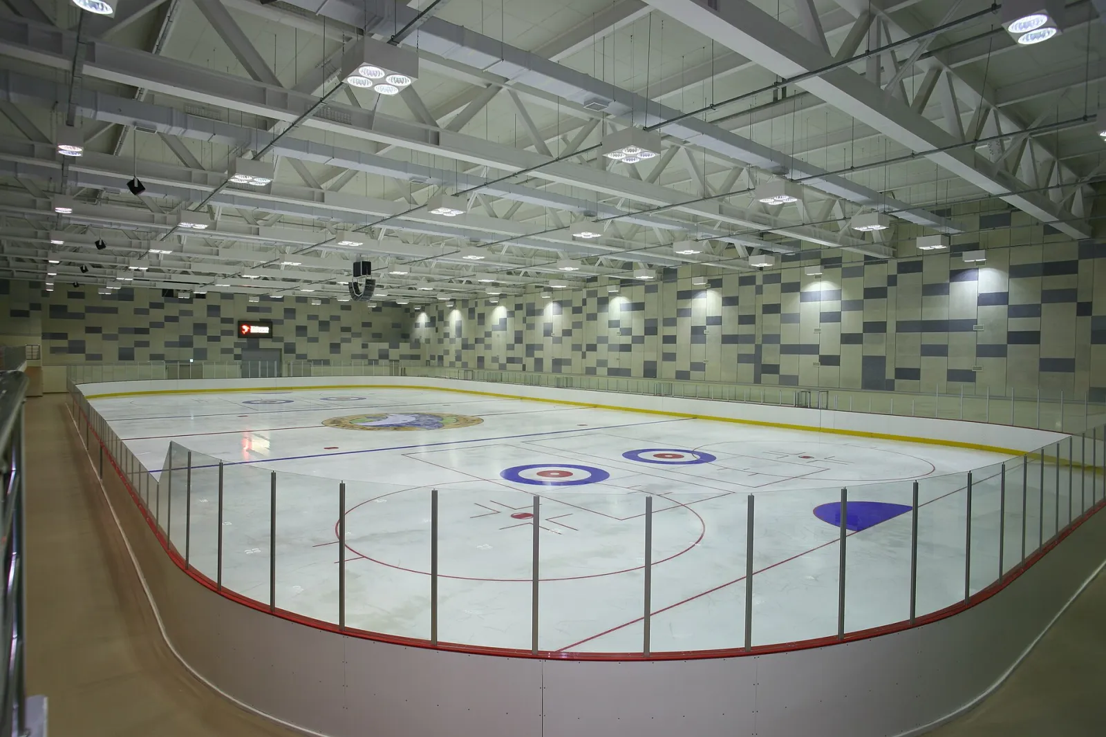 The empty ice of the Asan Gymnasium rink at Ulsan College, with curling circles and the faceoff markings painted under the steel-trussed ceiling, seen from the end of the stand. — Asan Gymnasium Ice Rink (Ulsan College)