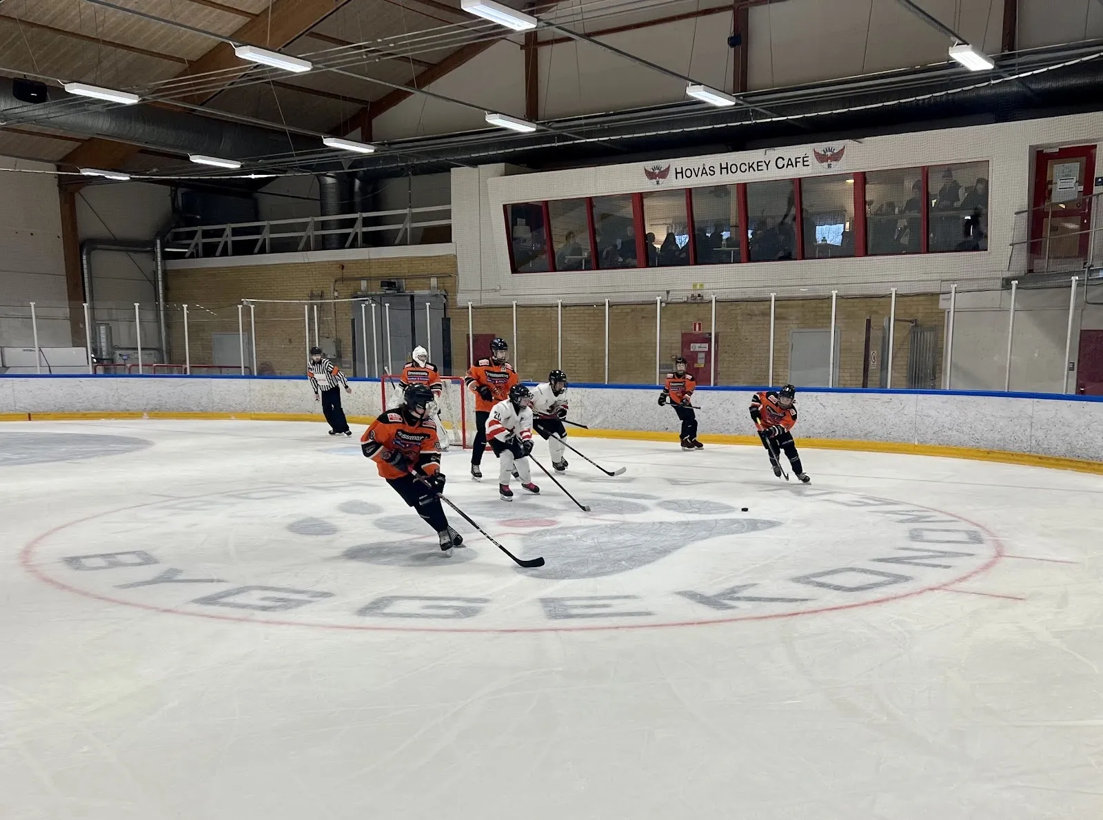 En ungdomsmatch i ishockey på isen i Askims ishall i Göteborg, sedd från sargen över hela isytan, med läktaren och taket bakom spelarna. – Askims ishall