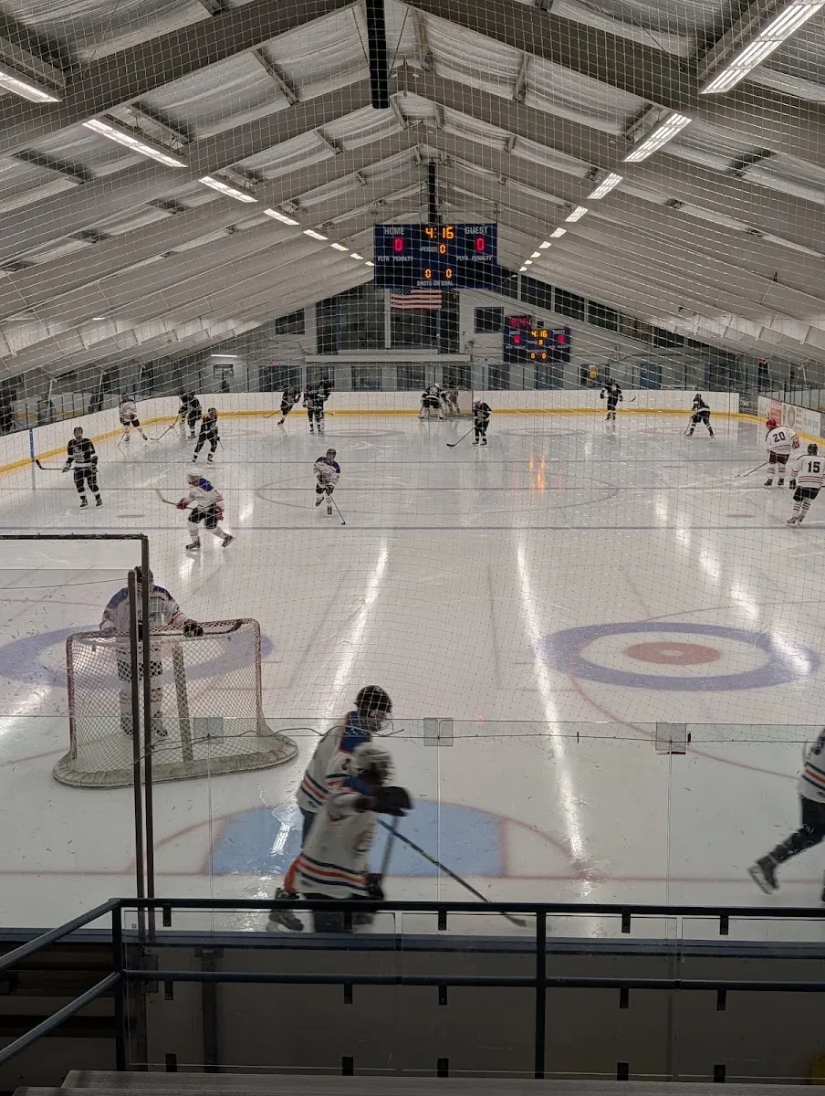 The rink during a hockey game, seen from the stands under the pitched wooden roof with the scoreboard at the far end. — Aspen Ice Garden