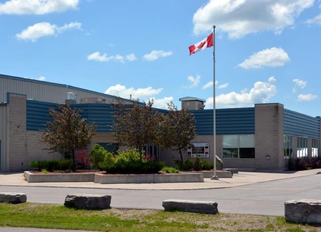 The community centre's low front with blue accent panels and a Canadian flag, behind landscaped beds by the parking lot. — Asphodel-Norwood Community Centre