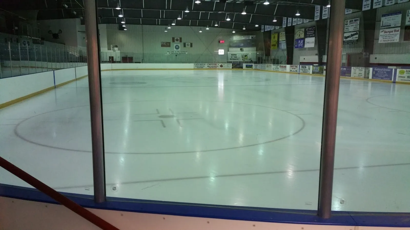 The empty ice sheet at Asphodel-Norwood Community Centre seen through the glass from rink level, with the centre-ice circle and the end wall's flags and sponsor boards beyond. — Asphodel-Norwood Community Centre