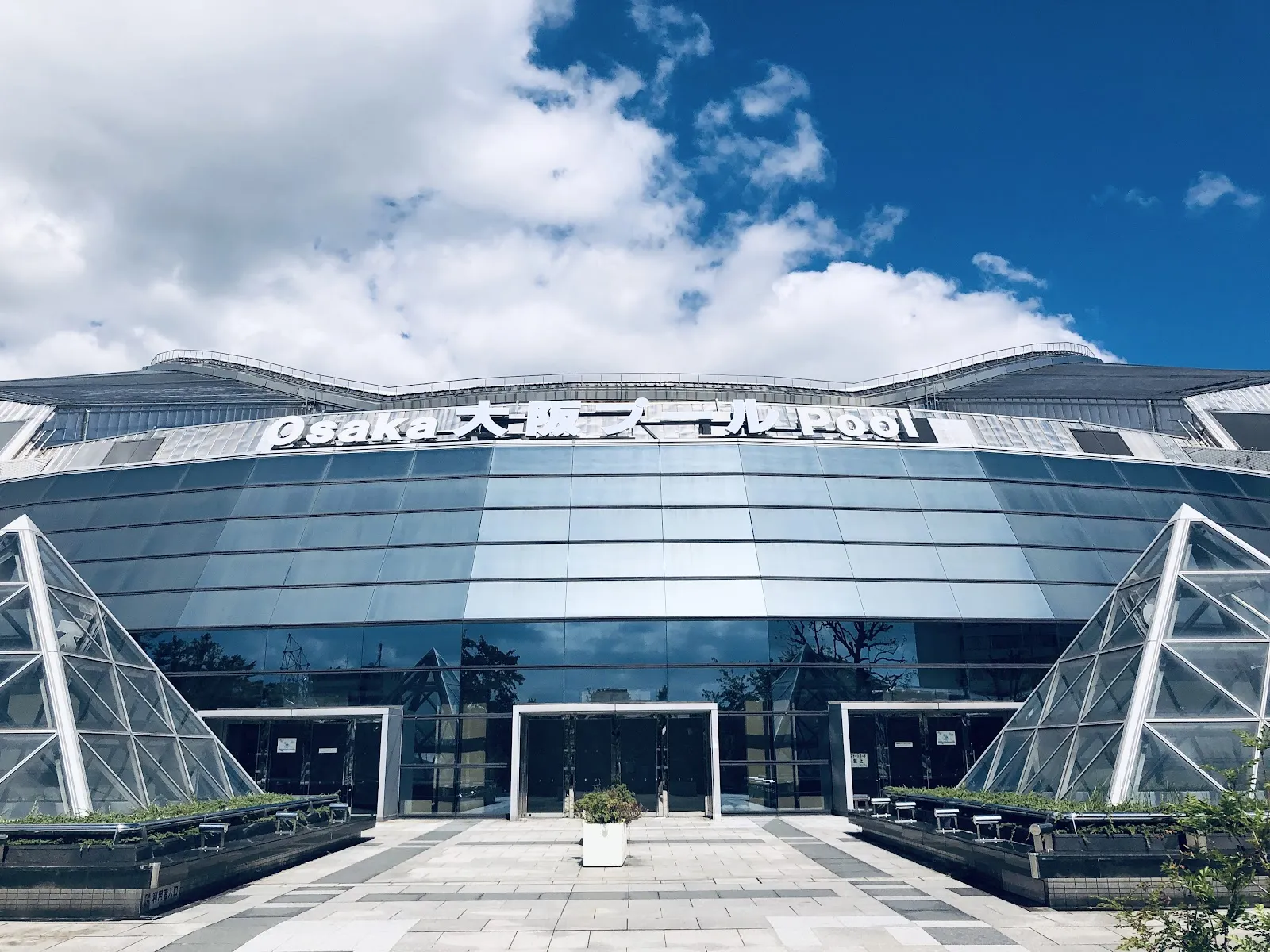 The glass-and-steel front of the Osaka Pool building in Yahataya Park, with its Osaka Pool sign over the doors and a paved plaza in front. — Asue Osaka Pool Ice Skating Rink