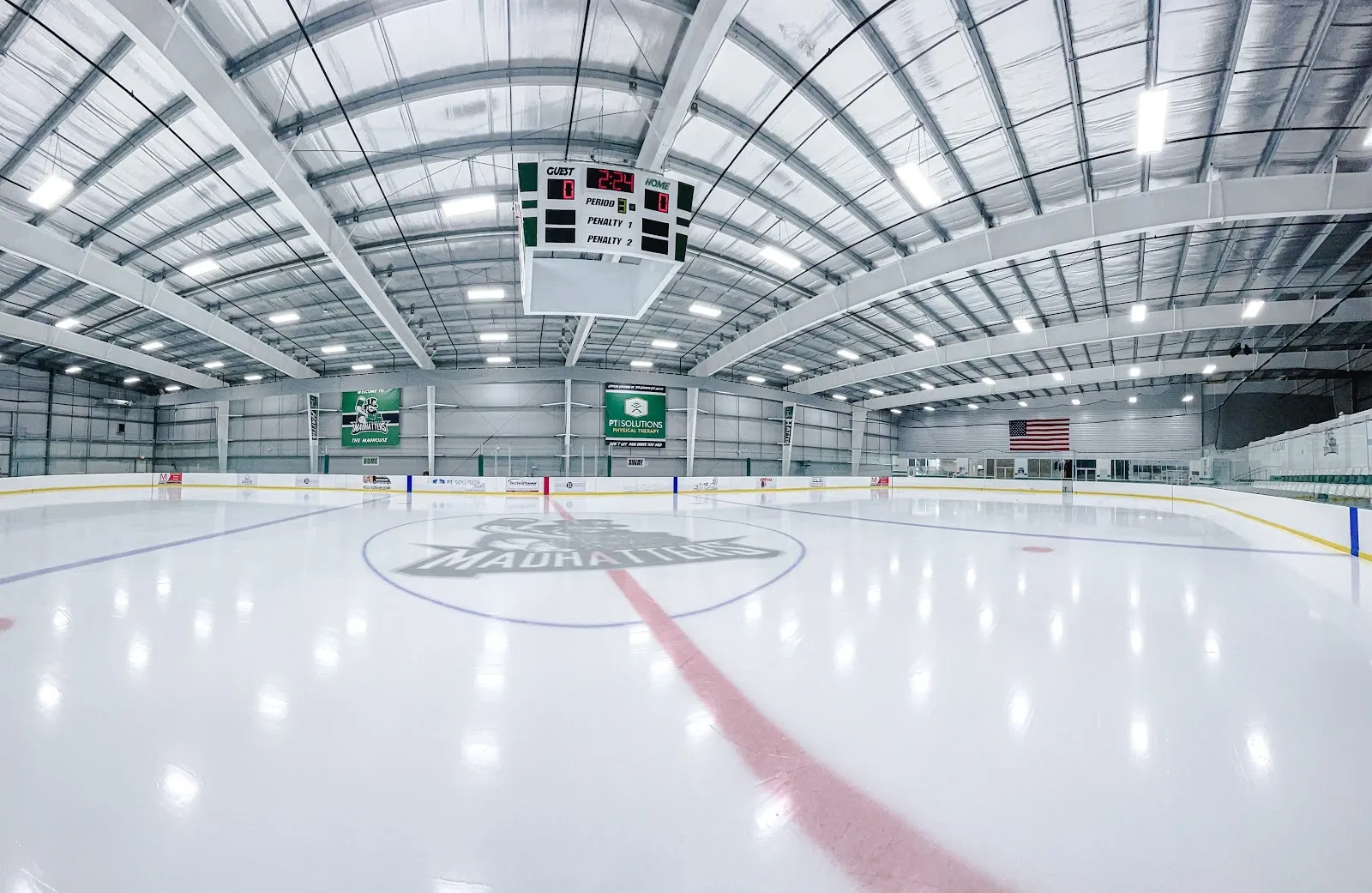 Una pista vacía vista desde las vallas del extremo, con hielo blanco, un logotipo en el centro y una línea roja, bajo un alto techo de acero arqueado con un marcador colgado sobre el extremo opuesto. — Atlanta Ice House