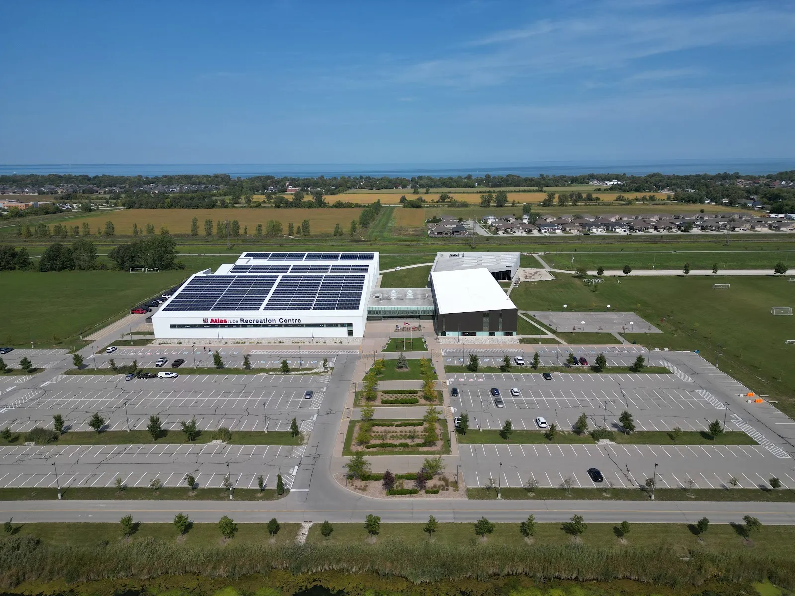 Vista aérea del Atlas Tube Recreation Centre, con su techo de paneles solares y su fachada con letrero detrás del gran estacionamiento, con campos y el lago St. Clair más allá. — Atlas Tube Recreation Centre