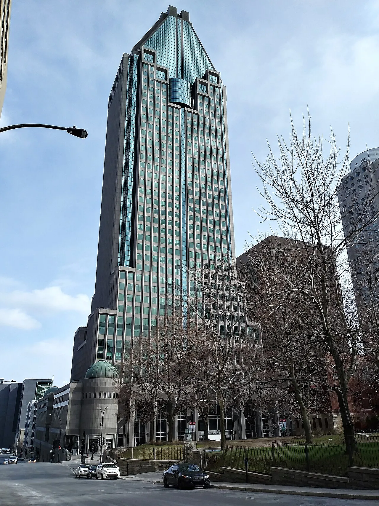The 1000 De La Gauchetière tower seen from street level, its stepped glass crown rising above the sidewalk trees; the rink occupies the building's ground-floor atrium. — Atrium Le 1000