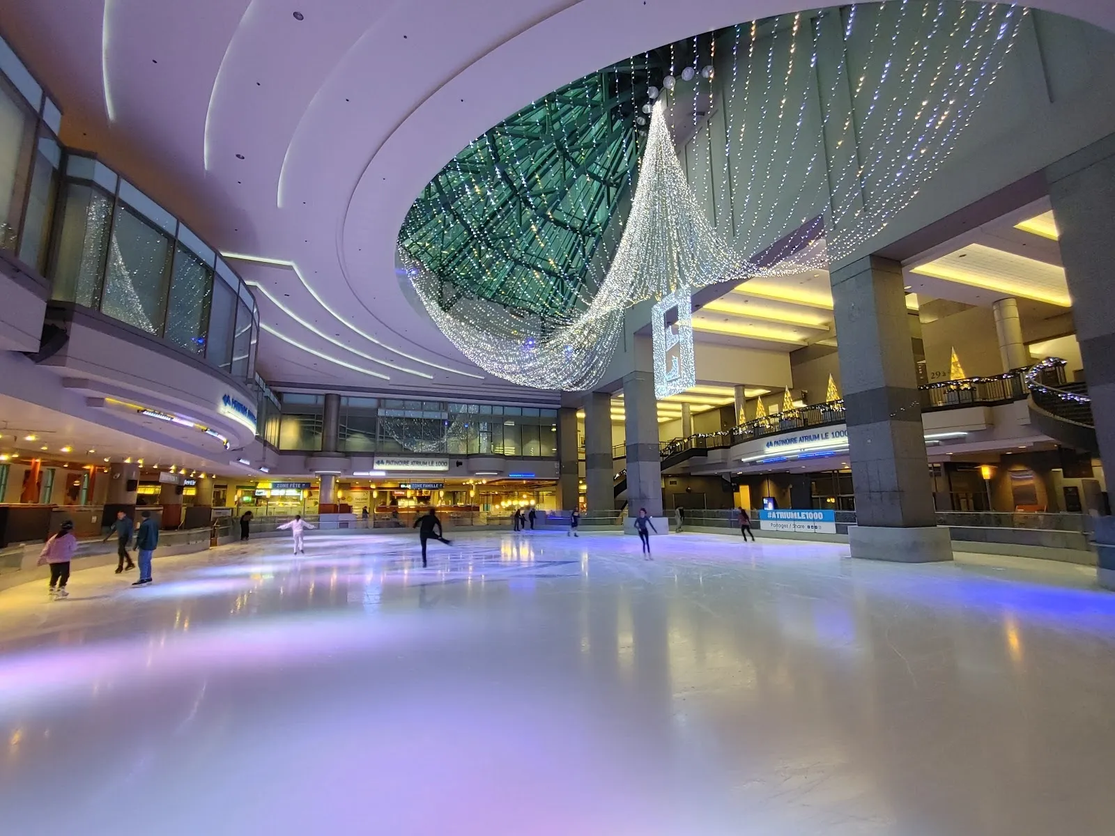 La pista bajo la cúpula de vidrio del atrio, con patinadores repartidos por el hielo y los balcones y las luces del centro comercial rodeando el piso. — Atrium Le 1000