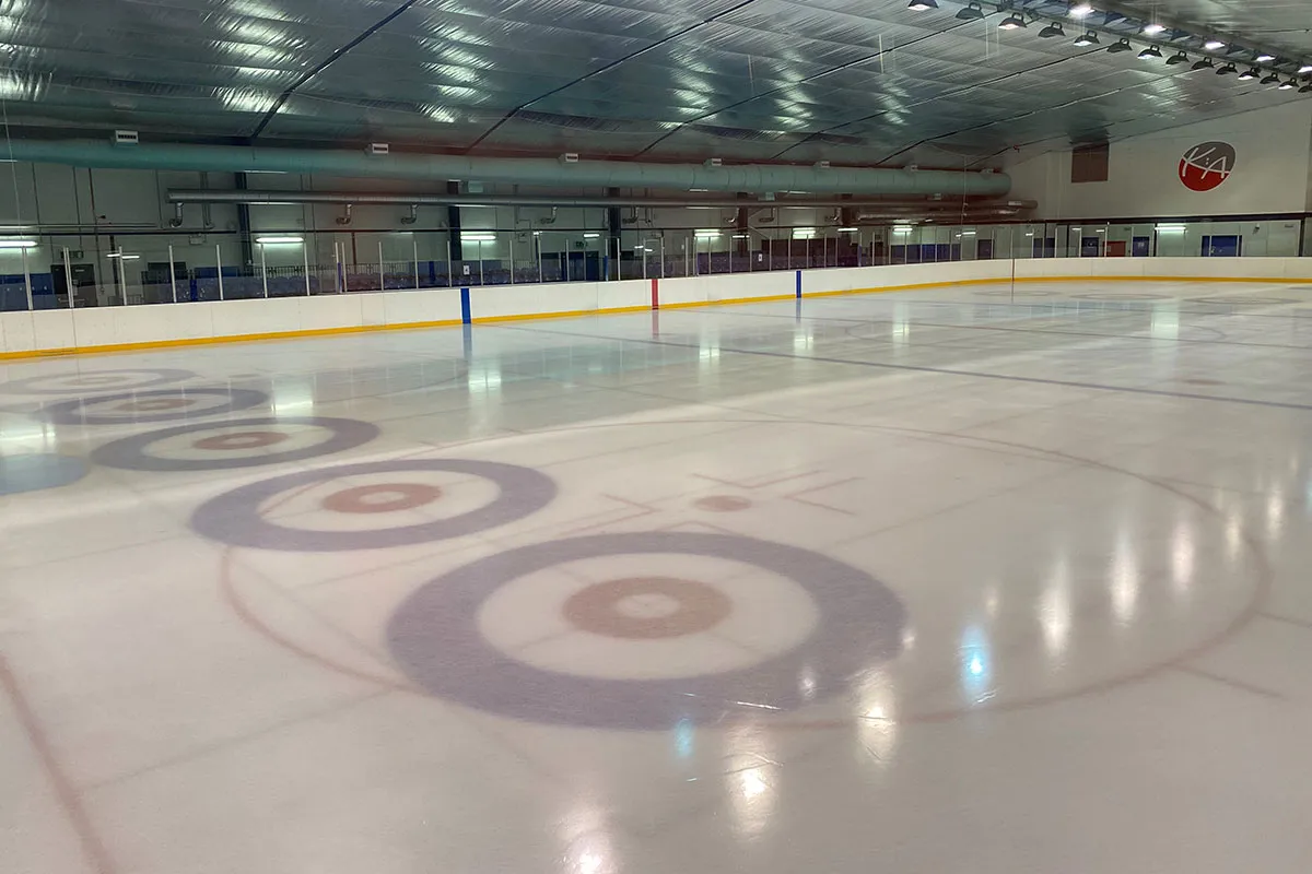 The empty ice from one corner, with curling rings painted on the surface and the boards and seating along the far side. — Auchenharvie Ice Rink